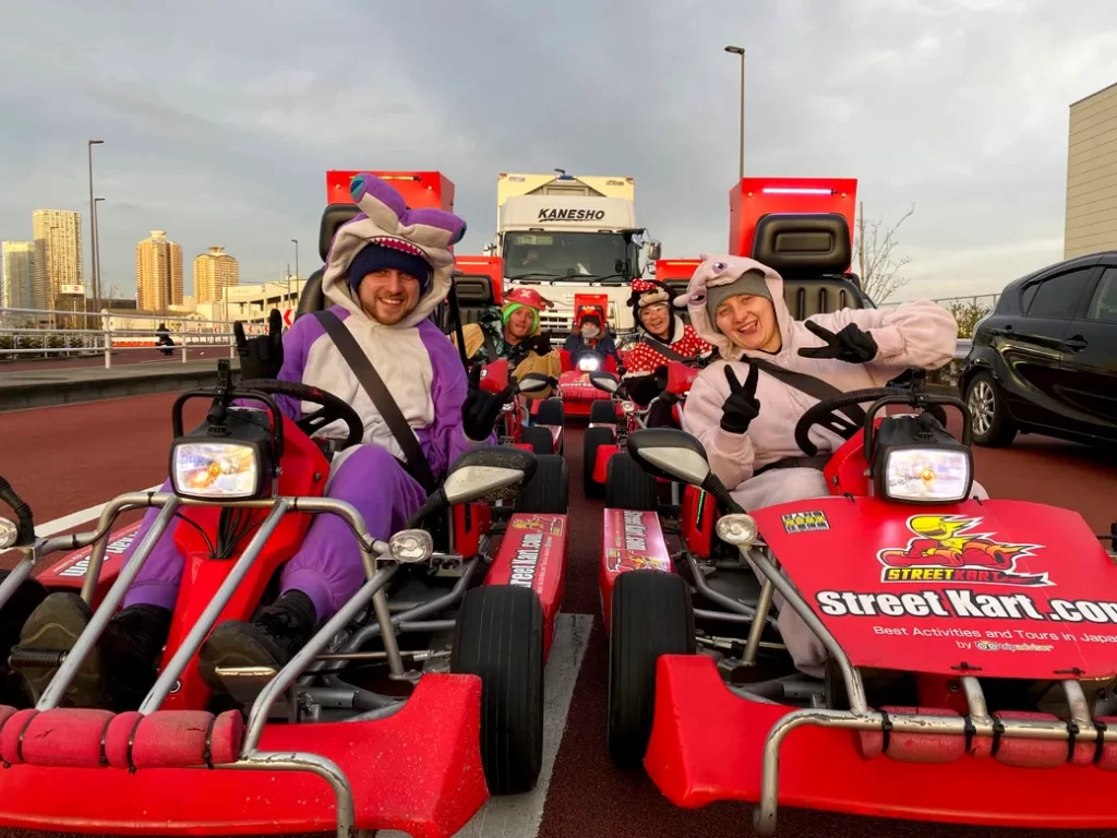 Group of people in colorful animal onesies driving red go-karts on a city street, posing and showing peace signs.