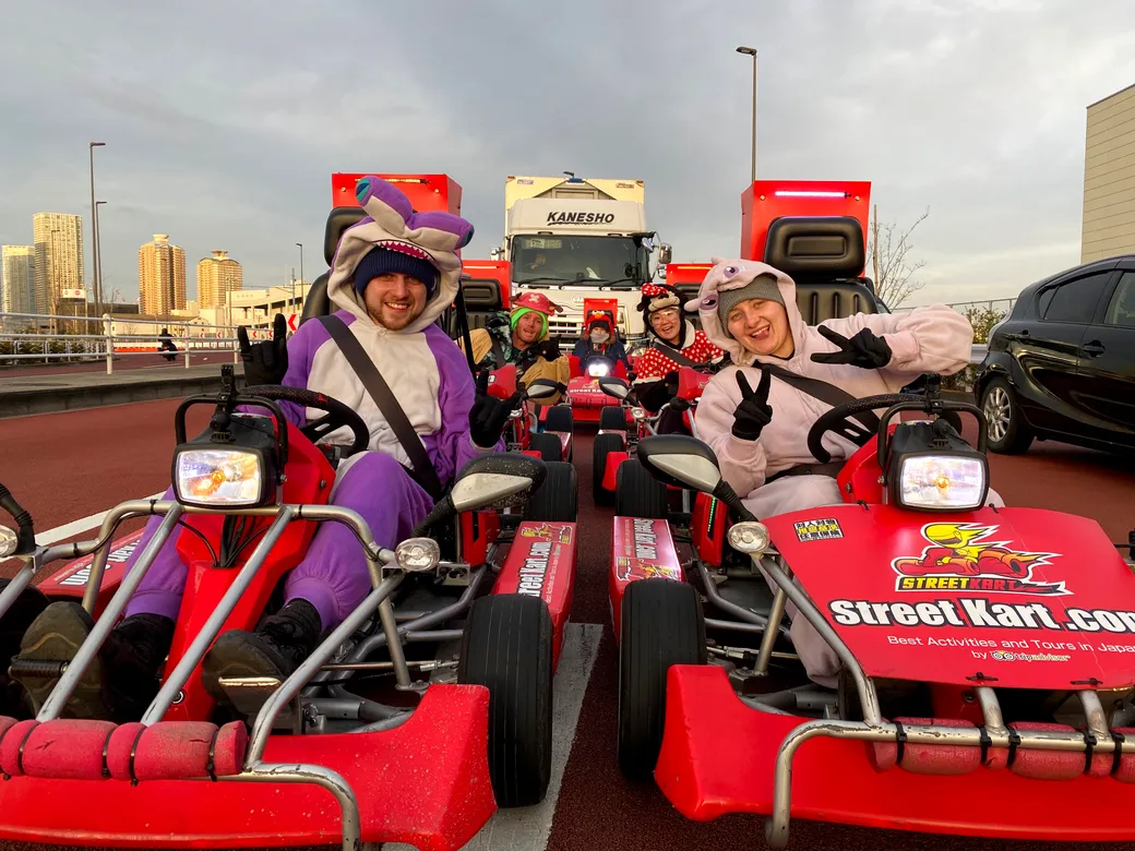 Group of people in colorful animal onesies driving red go-karts on a city street, posing and showing peace signs.