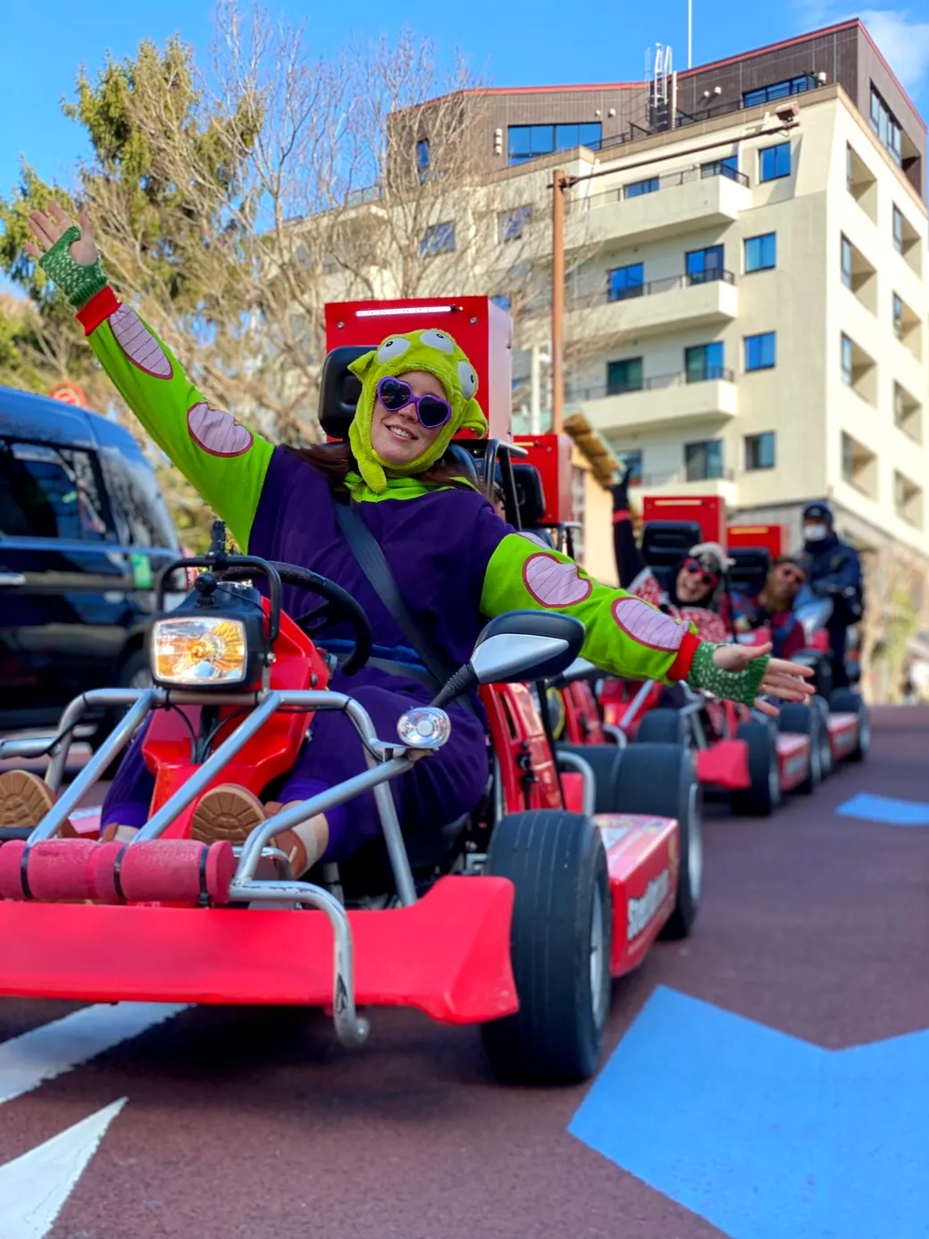 Front rider in a bright green and purple costume with a monster hat drives a red go-kart and waves in a city parade.