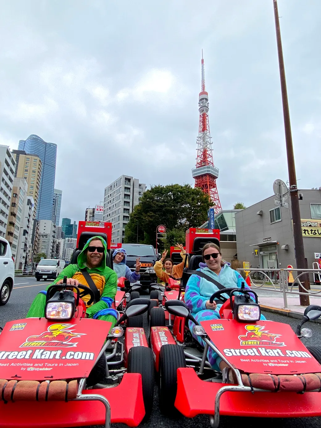 Group of people in bright costumes riding red go-karts down a city street with Tokyo Tower in the background, fun tour scene.