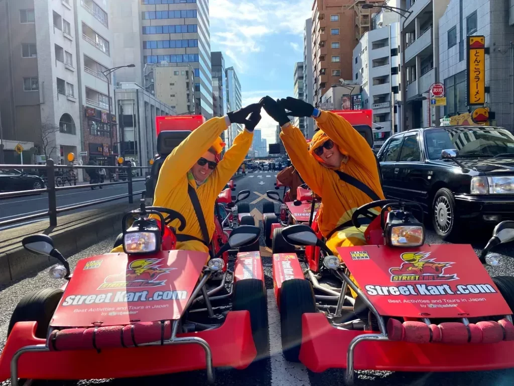 IMG_9188 - StreetKart Two people in yellow outfits form a heart shape with their arms while sitting in red Street Kart go-karts on a city street in Japan.